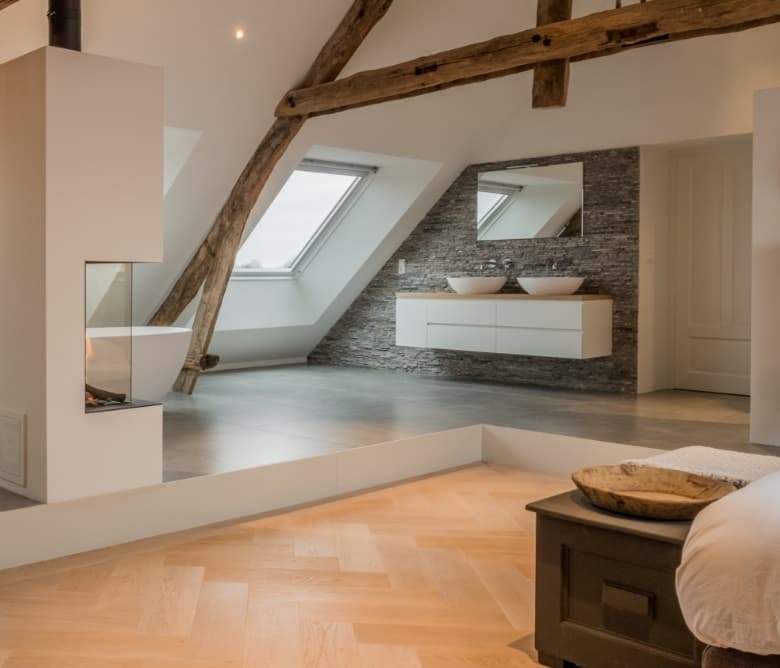 Badkamer op zolder onder oude balken, met een vrijstaand bad, visgraatvloer en een natuurstenen wand