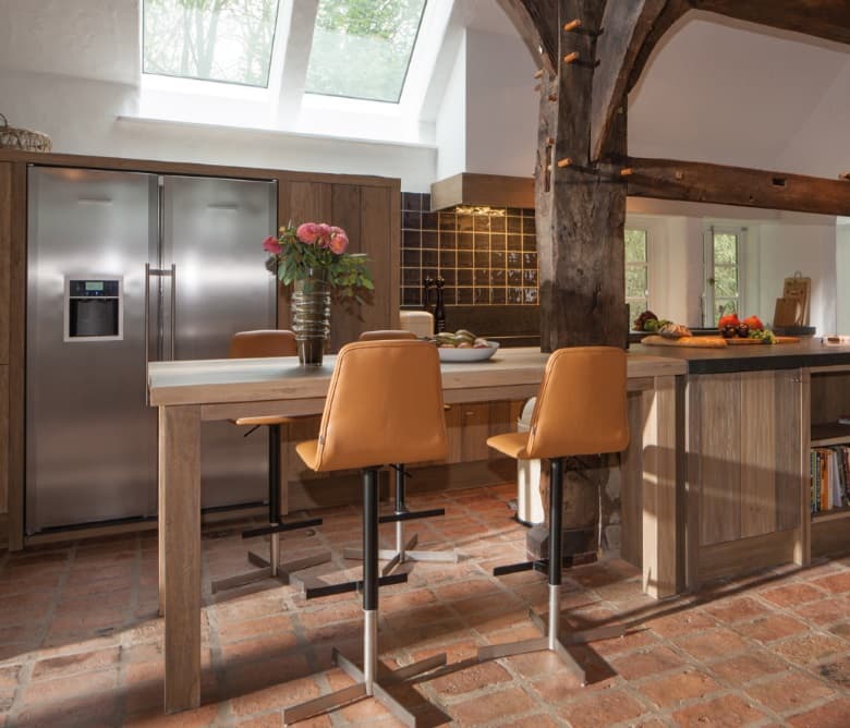 Keuken onder een open kapconstructie, met eiken maatwerk, een hoge bartafel met leren krukken en een rvs koelkast