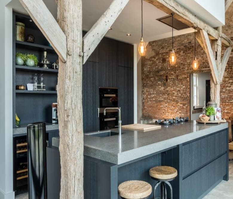 Donkere keuken met betonlook werkblad tegen een muur van oud metselwerk, met barkrukken en hanglampen